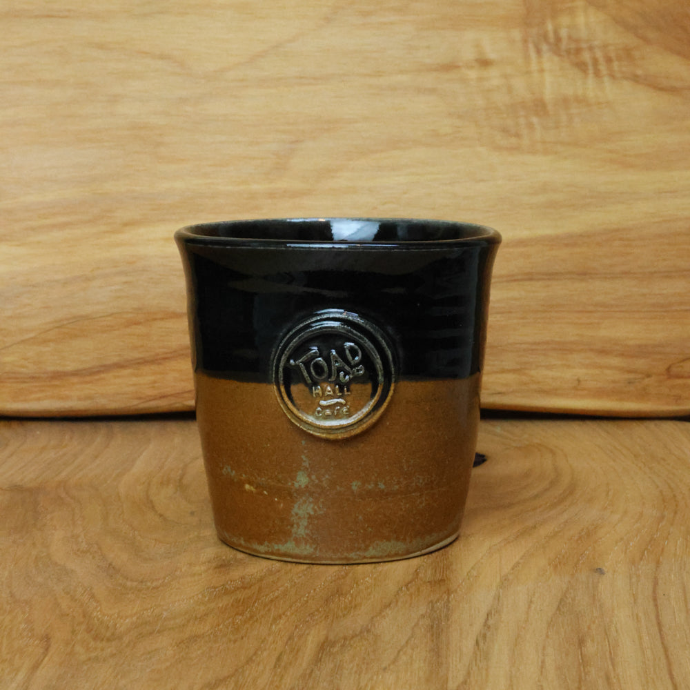 A handmade ceramic mug with a glossy black top half and a textured brown bottom half, featuring a raised circular 'TOAD HALL" logo on the front, set against a wooden background.