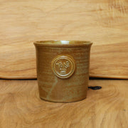 A small handmade ceramic cup with a brown and gold glaze, featuring an embossed circular emblem on the front, set against a wooden background.