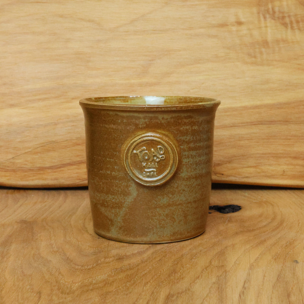 A small handmade ceramic cup with a brown and gold glaze, featuring an embossed circular emblem on the front, set against a wooden background.