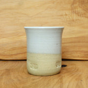 A handmade ceramic cup with a two-tone glaze, featuring a light gray upper half and a sandy beige lower half with two small leaf-shaped cutouts, set against a wooden background.