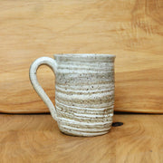 A handmade ceramic mug with a textured, speckled glaze in off-white and beige tones, set against a warm wood grain background.