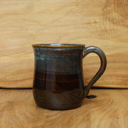A handmade ceramic mug with a dark brown and black glaze, featuring a curved handle and subtle floral relief details near the base, set against a light wood background.