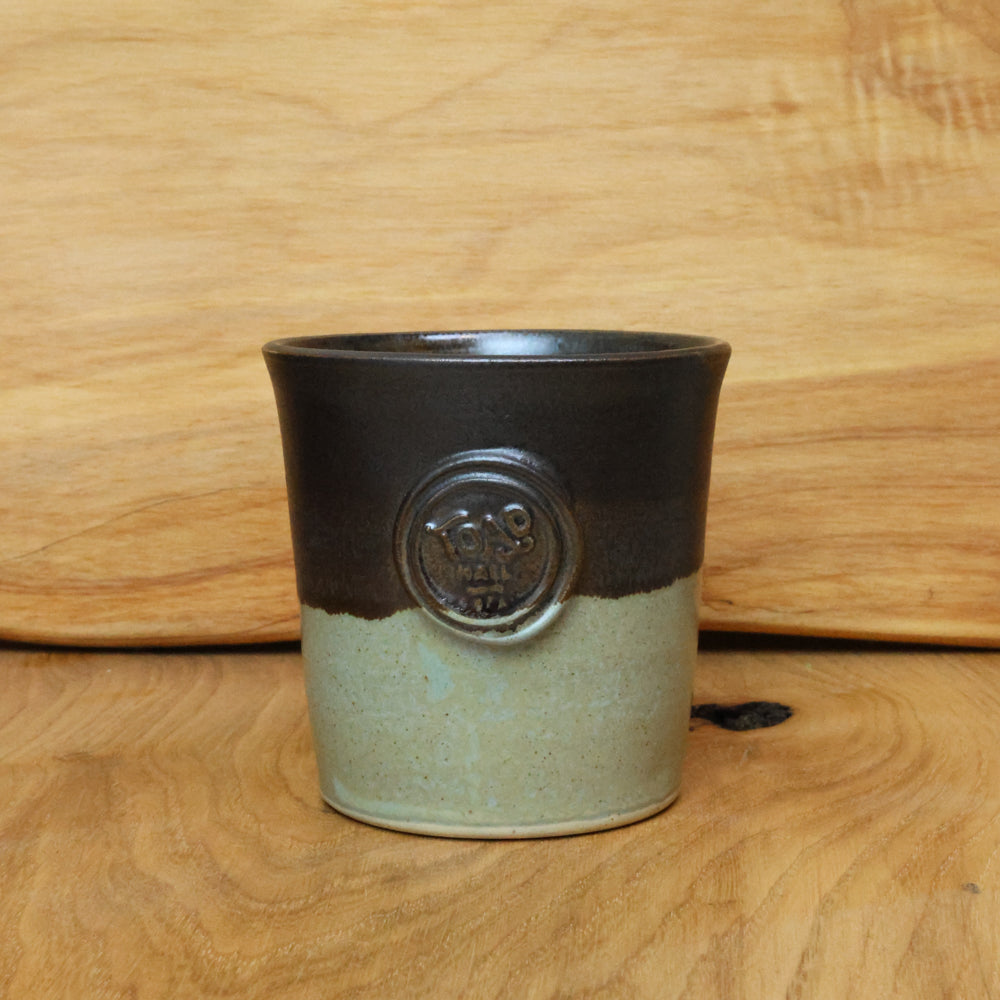 A handmade ceramic cup with a two-tone glaze, featuring a dark brown top half and a speckled light greenish-gray bottom half, set against a wooden background.